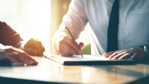 Person in business attire signing a document on a desk with sunlight coming through a window, while another person sits nearby.