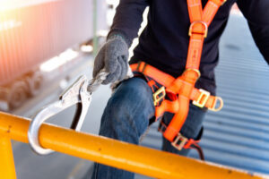 A person wearing a safety harness and gloves attaches a lanyard hook to a yellow railing while working on a corrugated metal roof.