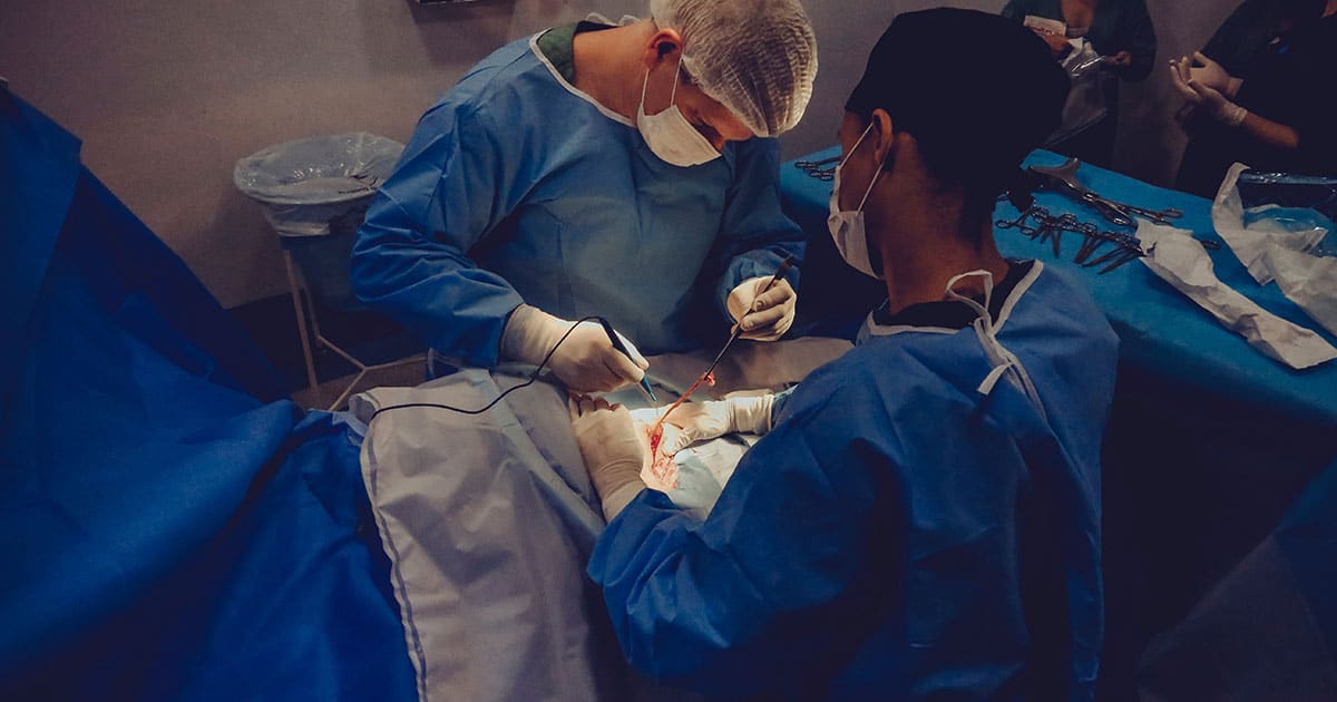 Two surgeons in blue surgical gowns and masks perform an operation on a patient in an operating room, using medical instruments under focused lighting.