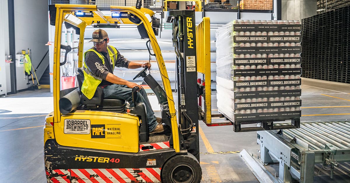 A worker operates a Hyster forklift to move a pallet of boxed goods inside a warehouse facility.