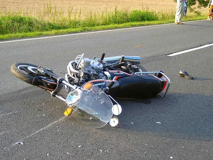 A motorcycle lies on its side on a paved road, with a single shoe nearby. Grass and a field are visible in the background.