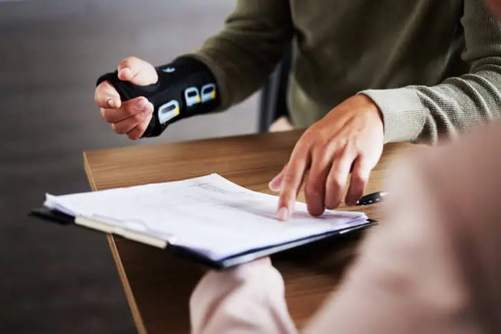 Two people sit at a table; one wears a wrist brace and points to documents on a clipboard while the other person listens.