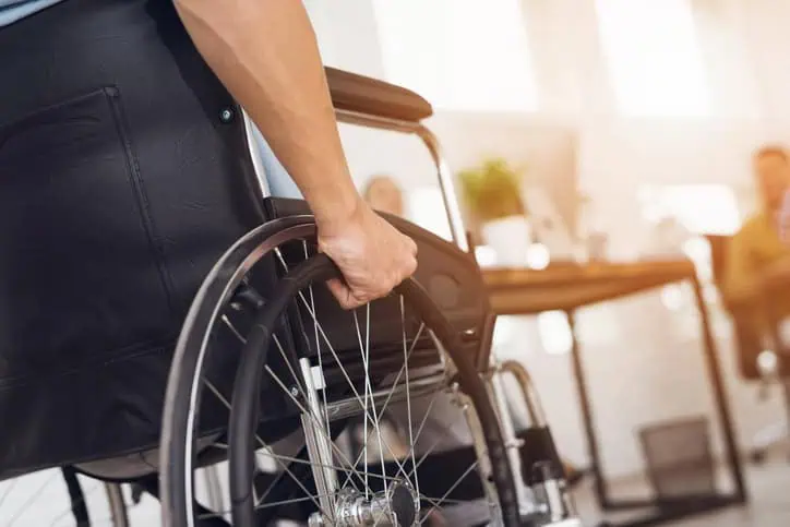 A person’s hand grips the wheel of a wheelchair in an indoor office setting with sunlight streaming in.