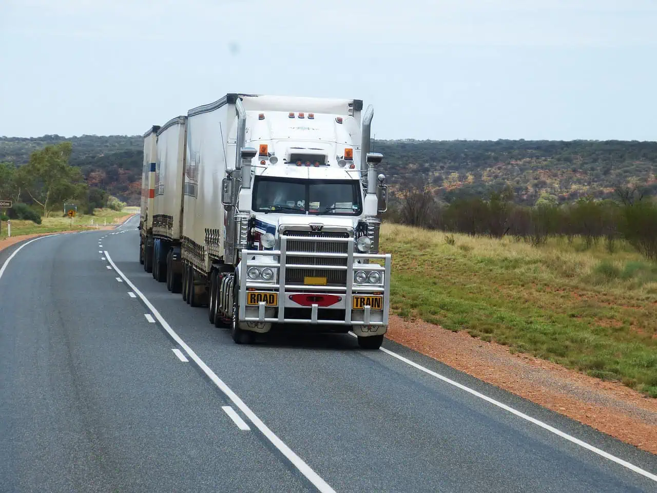 A large white road train truck with two trailers drives along a two-lane highway through a rural, grassy landscape under a partly cloudy sky.
