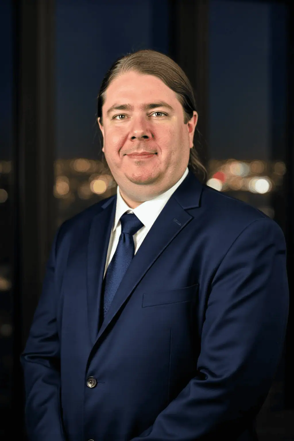 A man with long brown hair in a dark blue suit and tie stands indoors in front of windows showing a blurred cityscape at night.