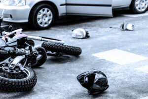 A motorcycle lies on its side near a crosswalk, with helmets and debris scattered on the road beside a car.