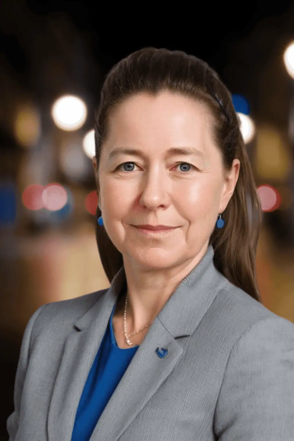 A woman in a gray blazer and blue top stands in front of a blurred night cityscape with bokeh lights.