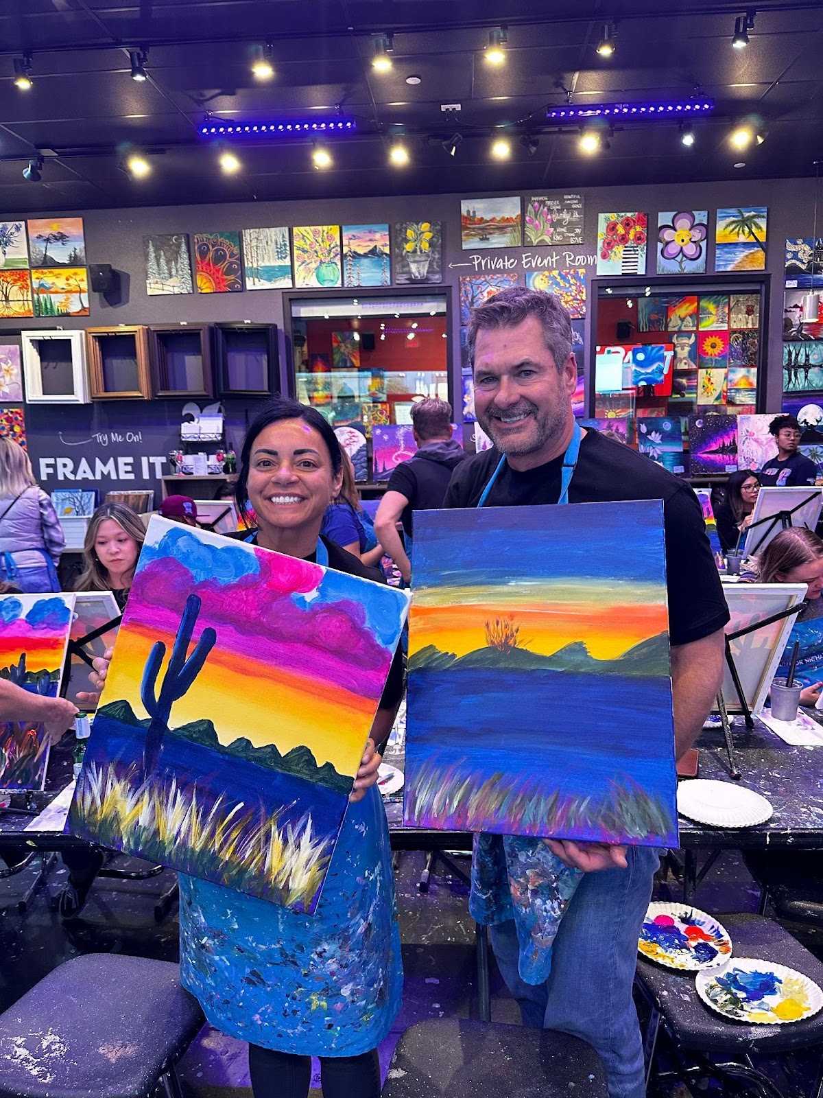 a woman and a man holding up paintings of desert landscapes