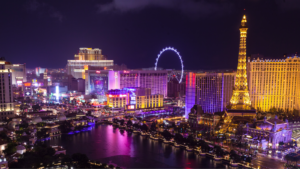 Night view of the Las Vegas Strip with illuminated hotels, casinos, the Eiffel Tower replica, and the High Roller observation wheel reflected in water.