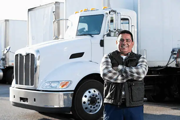 A man in a plaid shirt and vest stands with arms crossed, smiling in front of a white semi-truck in a parking lot.