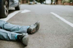 A person in dress pants and black shoes is lying on their back on a paved street, partially visible from the waist down. A car and road markings are nearby.