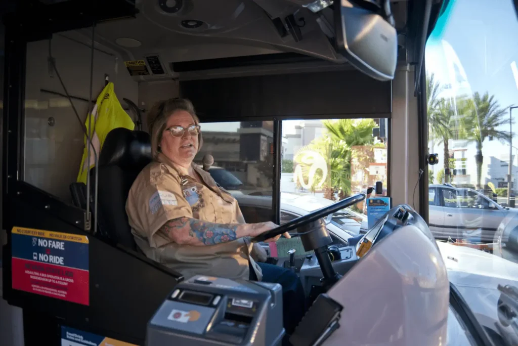 Un conductor de autobús con gafas y tatuajes está sentado en el asiento del conductor de un autobús urbano, mirando hacia adelante. Se ven señales, una chaqueta de alta visibilidad y un paisaje urbano.