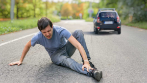 A man sits on a road clutching his ankle in pain as a car drives away in the background.