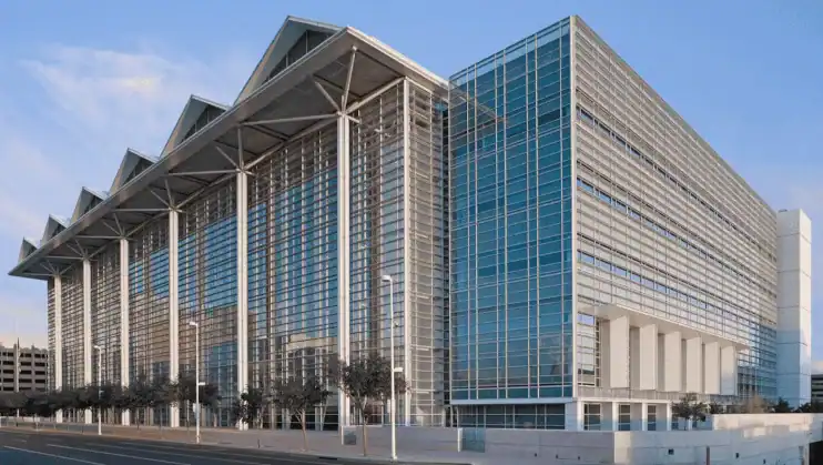Large modern glass and steel building with a high, peaked roof and rows of vertical supports along the facade, located on a city street.