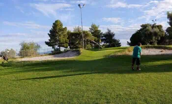 A child in a green shirt stands on a grassy field with trees and a light pole in the background under a partly cloudy sky.