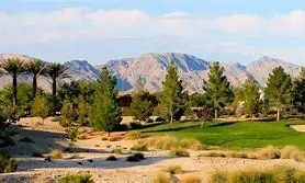 A desert golf course with green fairways, scattered trees, and mountains in the background under a partly cloudy sky.