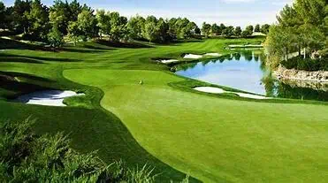 A well-maintained golf course with green fairways, sand bunkers, trees, and a water hazard under a clear sky.