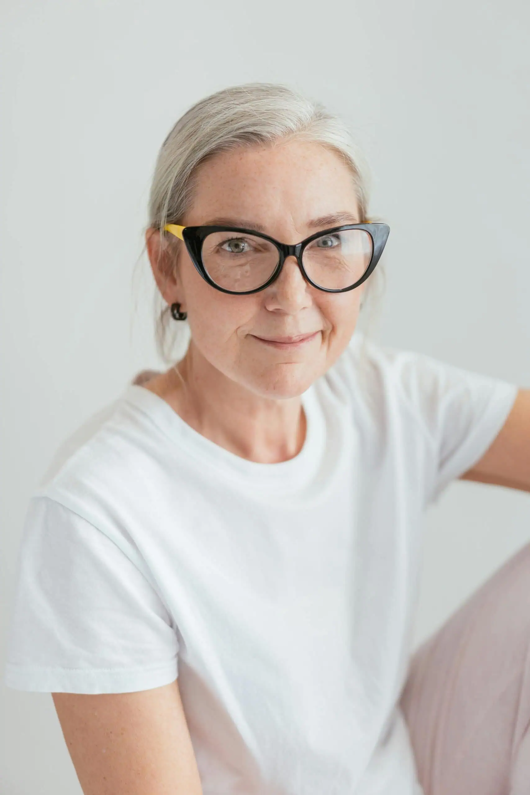 Older woman with gray hair, wearing black cat-eye glasses and a white t-shirt, sitting against a plain light background and looking at the camera.