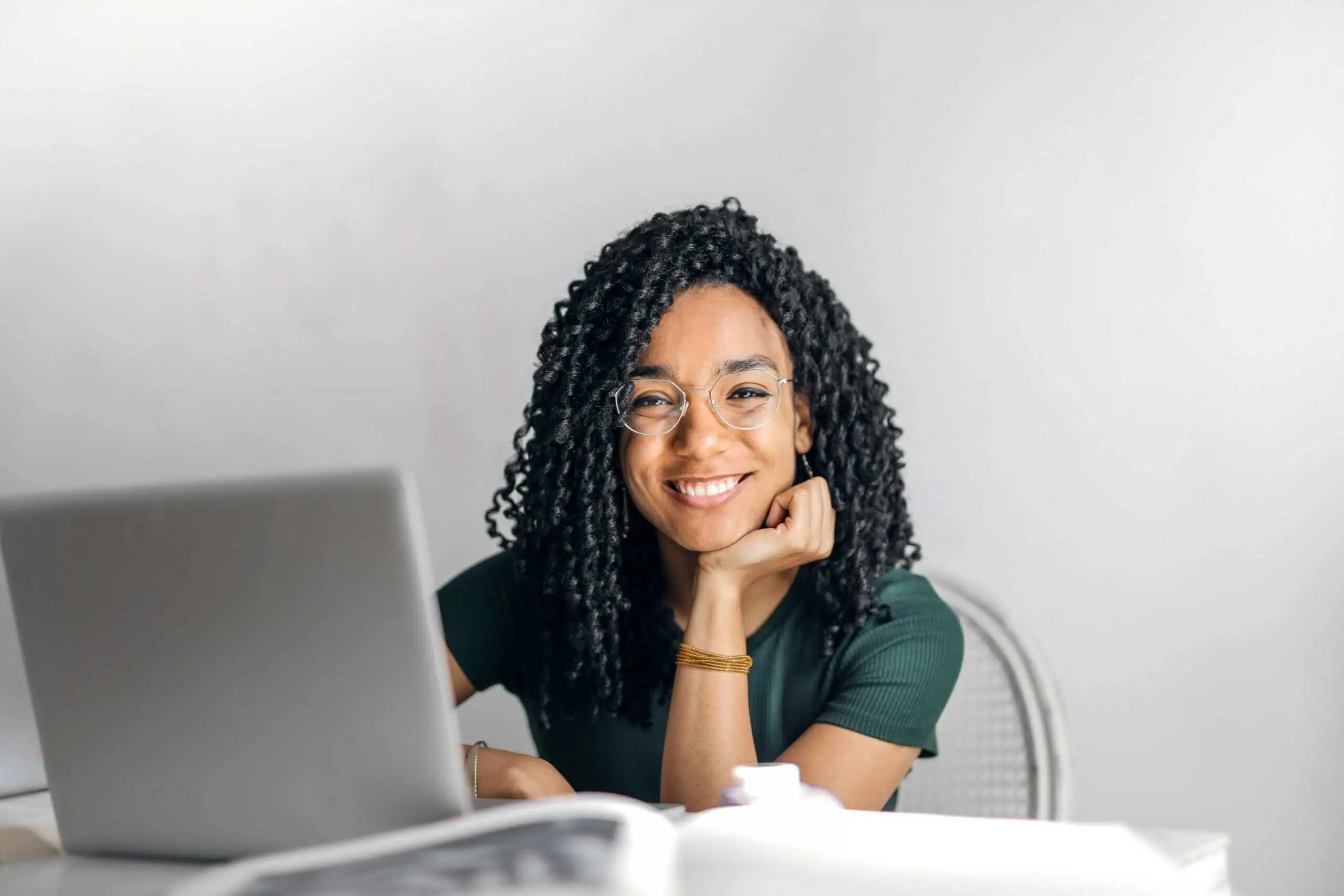 A person with curly hair and glasses, wearing a green shirt, sits at a desk smiling, with an open laptop and books in front of them.