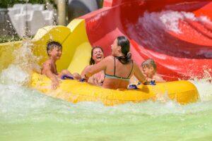 Four people ride a yellow inflatable raft down a water slide, splashing into a pool and smiling.