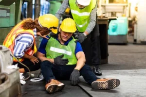 A worker wearing safety gear sits on the floor holding his abdomen, while two colleagues check on him in an industrial setting.
