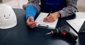 Person with a blue arm cast filling out a form on a clipboard at a desk with a safety helmet, laptop, and power drill nearby.