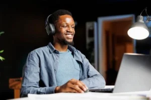 A man wearing headphones sits at a desk, smiling while looking at a laptop screen under a desk lamp.