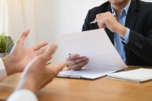 Two people sit at a desk during a meeting; one holds a document and pen while the other gestures with open hands.
