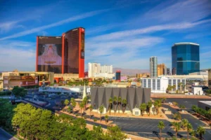 A cityscape view of Las Vegas featuring tall hotels, a prominent large screen display, palm trees, and a modern angular building in the foreground under a blue sky.