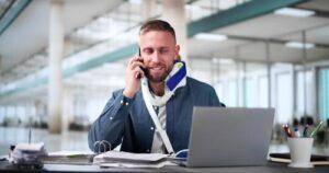 A man wearing a neck brace sits at a desk, talking on the phone while working on a laptop in a modern office setting.