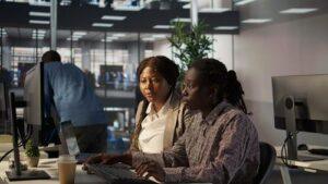 Two women working together at a computer in a modern office, with another person in the background and desks with monitors and plants around them.