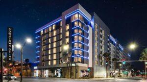 A multi-story hotel building with blue accent lighting on the exterior, located at a street corner at night, with a sign reading "Downtown Grand.