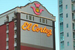 The exterior of the El Cortez building displays a bright, illuminated sign with its name and a decorative emblem above it, set against a clear sky.