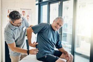 Physiotherapist examines an older man's lower back as he sits on an exam table in a clinic.