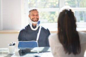 Man wearing a neck brace and arm sling sits across from a woman at a desk in a brightly lit room.