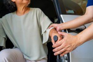 A person helps an older adult get out of a car by supporting their arm and using a mobility aid.