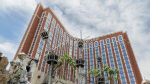 A tall, red-brick building with "Treasure Island" signage, pirate ship masts, palm trees, and statues in front under a partly cloudy sky.