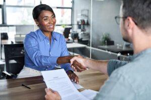 A woman in business attire smiles and shakes hands with a man holding a contract at a desk in a modern office.
