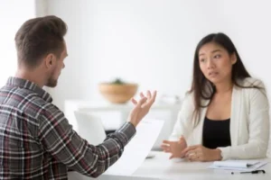 Two people sit across from each other at a desk, having a serious conversation. One holds a sheet of paper while the other listens with a concerned expression.