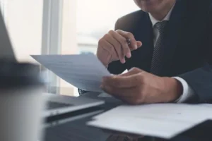 A person in a suit reviews documents at a desk with a laptop and papers in an office setting.