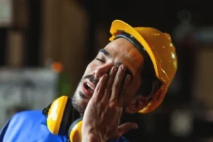 A worker wearing a yellow hard hat and protective headphones touches his face and appears tired or fatigued.