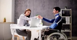 A woman and a man in a wheelchair with a neck brace sit at a desk, smiling and fist bumping across a laptop in a modern office setting.
