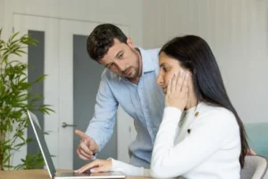 A man points at a laptop screen while a woman sits and looks at the screen with a concerned expression.