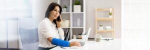 Woman with a blue arm cast sits at a desk, talking on the phone and working on a laptop in a bright, modern office.