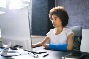 A woman with a blue cast on her right arm works at a computer desk in a modern office setting.