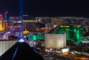 A nighttime aerial view of the Las Vegas Strip shows illuminated hotels and casinos, including the MGM Grand and the Luxor pyramid, against a background of city lights.