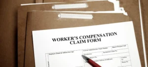 A close-up of a worker’s compensation claim form clipped to a brown file folder, with other folders stacked behind it and a red pen resting on the form.