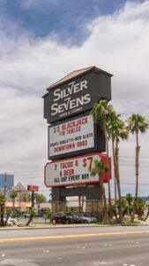 Silver Sevens Hotel & Casino sign displays promotions for blackjack, tacos, beer, and gaming, with palm trees and a cityscape in the background.