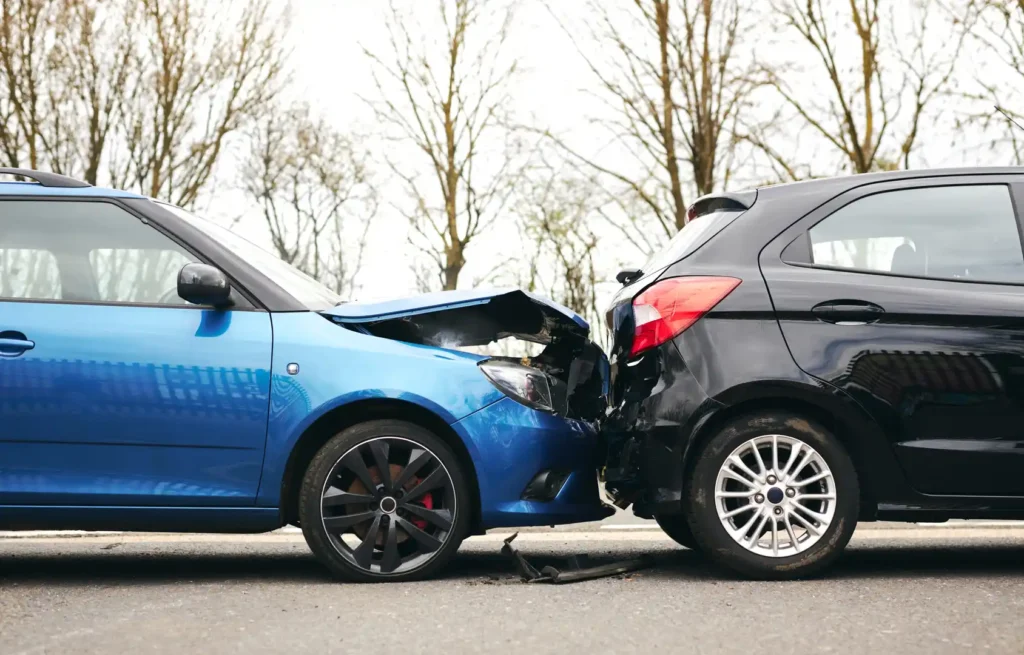 A blue car has rear-ended a black car on a road, causing visible damage to the front of the blue car and the rear of the black car. Trees are in the background.