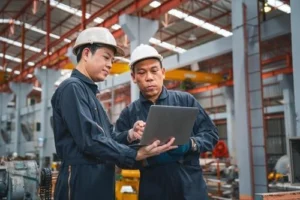 Two workers wearing hard hats and coveralls look at a laptop together inside a large industrial facility or factory.